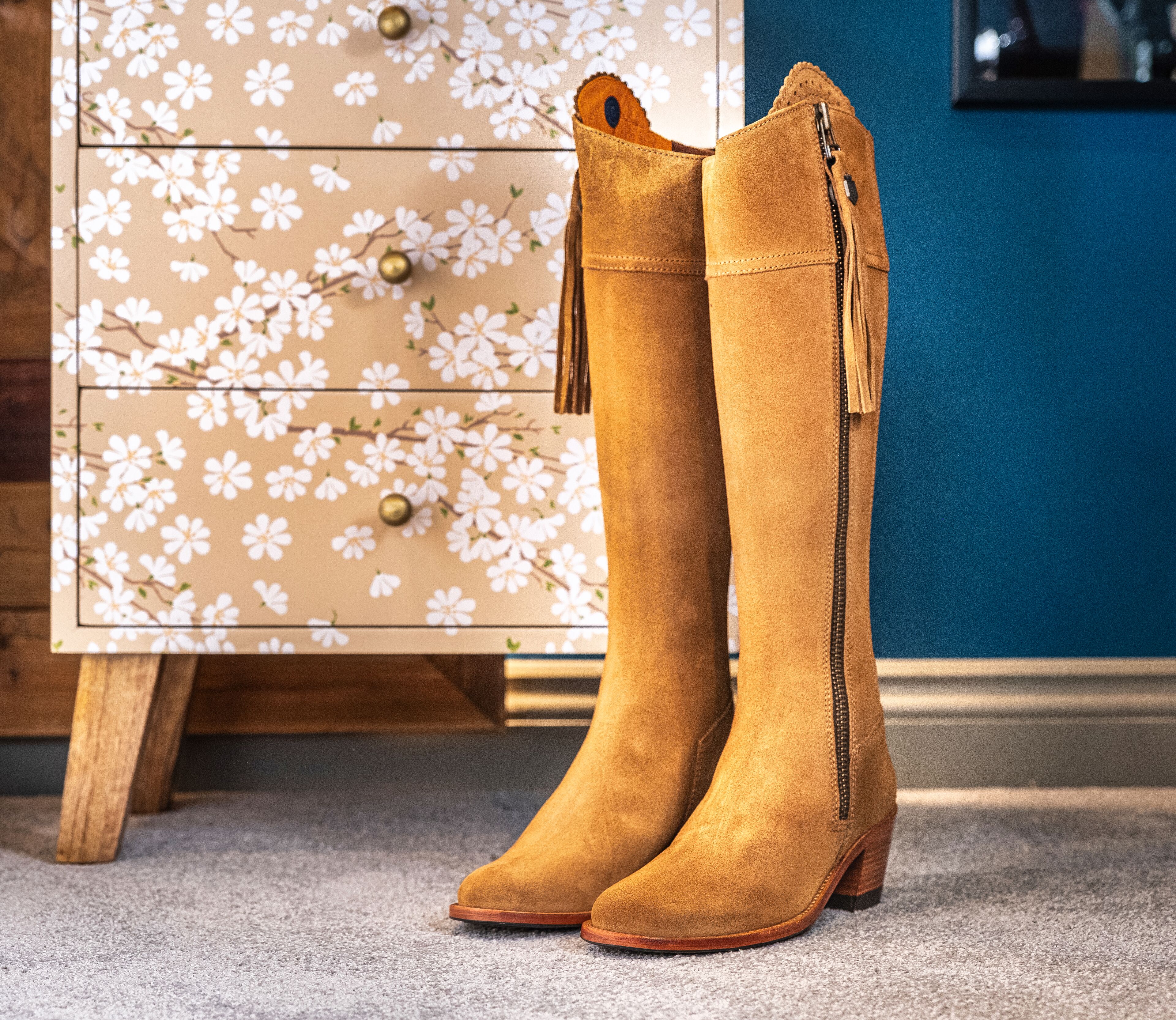 A pair of tan suede knee-high boots with a zipper and tassel detail, standing on a carpeted floor.