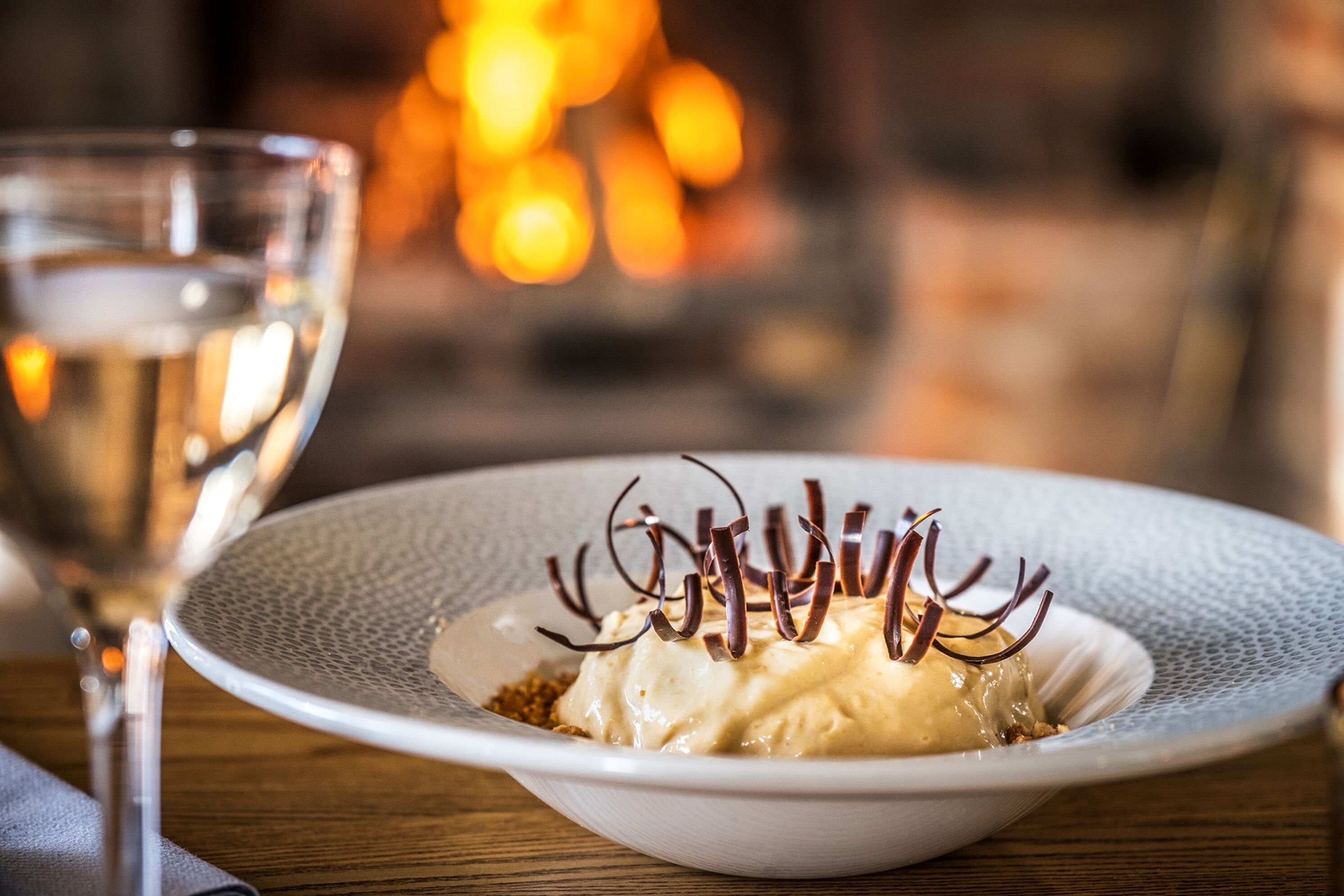 A dessert on a white plate with chocolate decorations, accompanied by a glass of white wine.