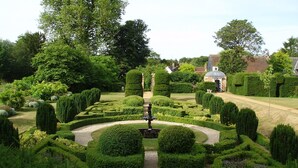 A meticulously landscaped garden with a central fountain, surrounded by geometrically trimmed hedges and various shrubs. - The Cricketers (Saffron Walden)