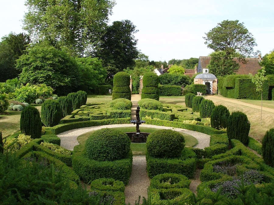 A meticulously landscaped garden with a central fountain, surrounded by geometrically trimmed hedges and various shrubs.