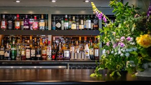A bar area with shelves of various liquor bottles, a bar counter, and a potted plant with flowers. - The Cricketers (Saffron Walden)
