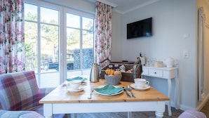 A dining area with a wooden table set for two, a wicker basket with bread, a coffee pot, and a white cabinet with a television above. - Hotel Tweed (St. Peter-Ording)