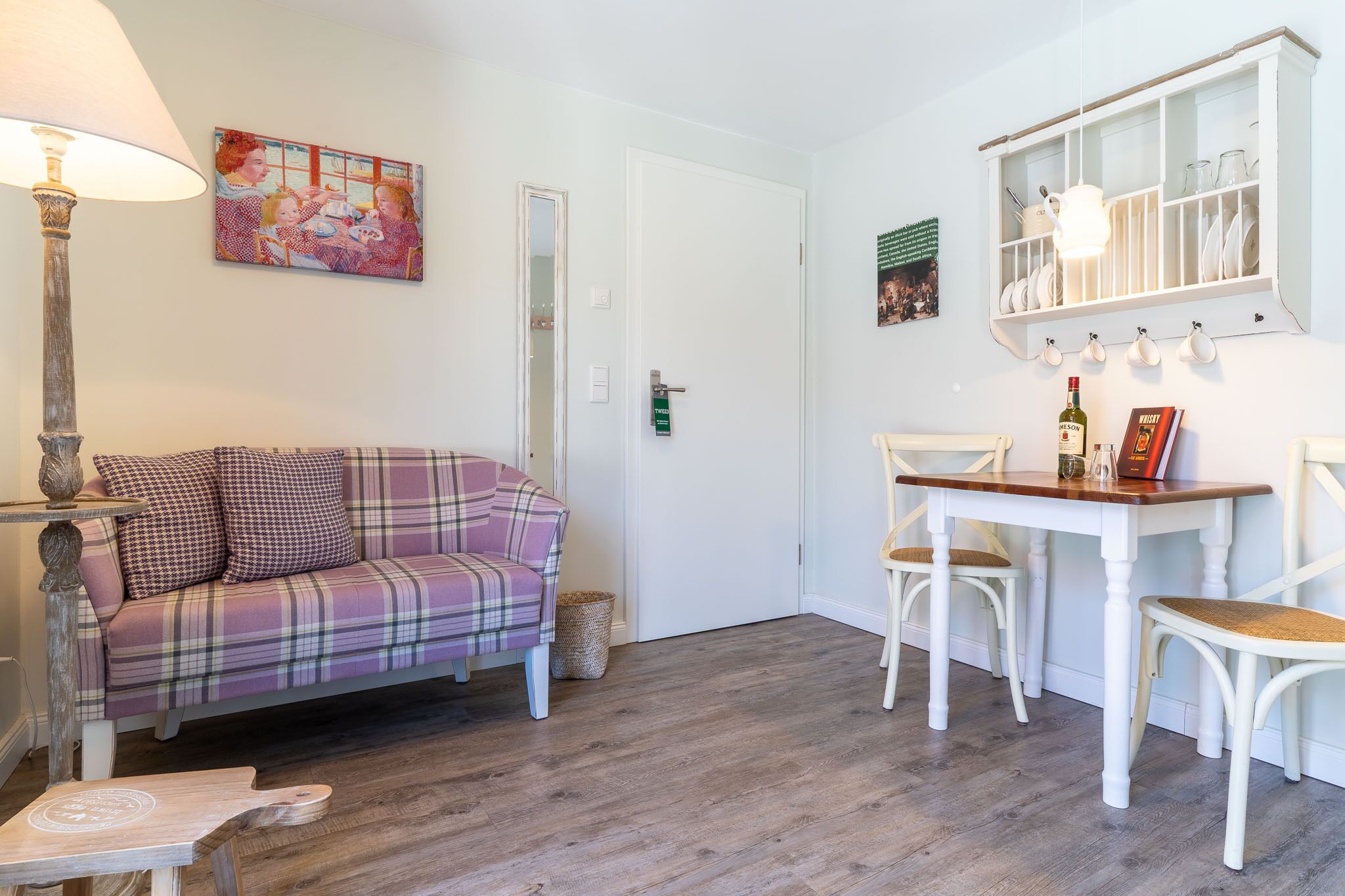 A cozy living room with a plaid sofa, a dining table with chairs, a lamp, and a wall-mounted cabinet with dishes.