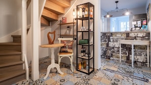 A staircase with wooden steps and white walls, a white chair with a patterned cushion, a black shelving unit with bottles and a green bag, and a tiled floor with a geometric pattern. - Hotel Tweed (St. Peter-Ording)