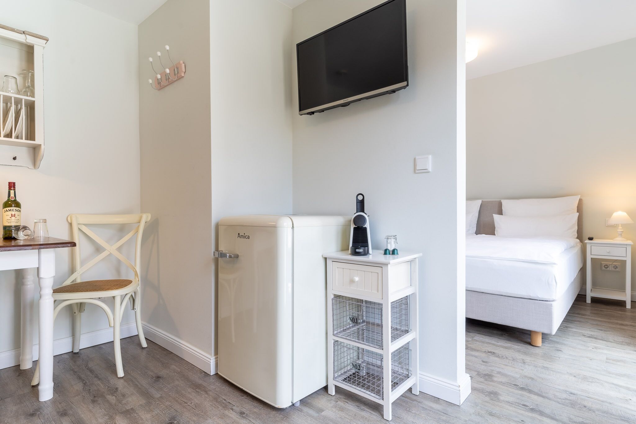 Habitación compacta con cama, refrigerador blanco pequeño, escritorio con silla, televisor montado en la pared y mesita de noche con lámpara.