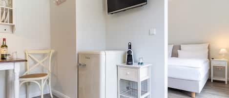 Habitación compacta con cama, refrigerador blanco pequeño, escritorio con silla, televisor montado en la pared y mesita de noche con lámpara.