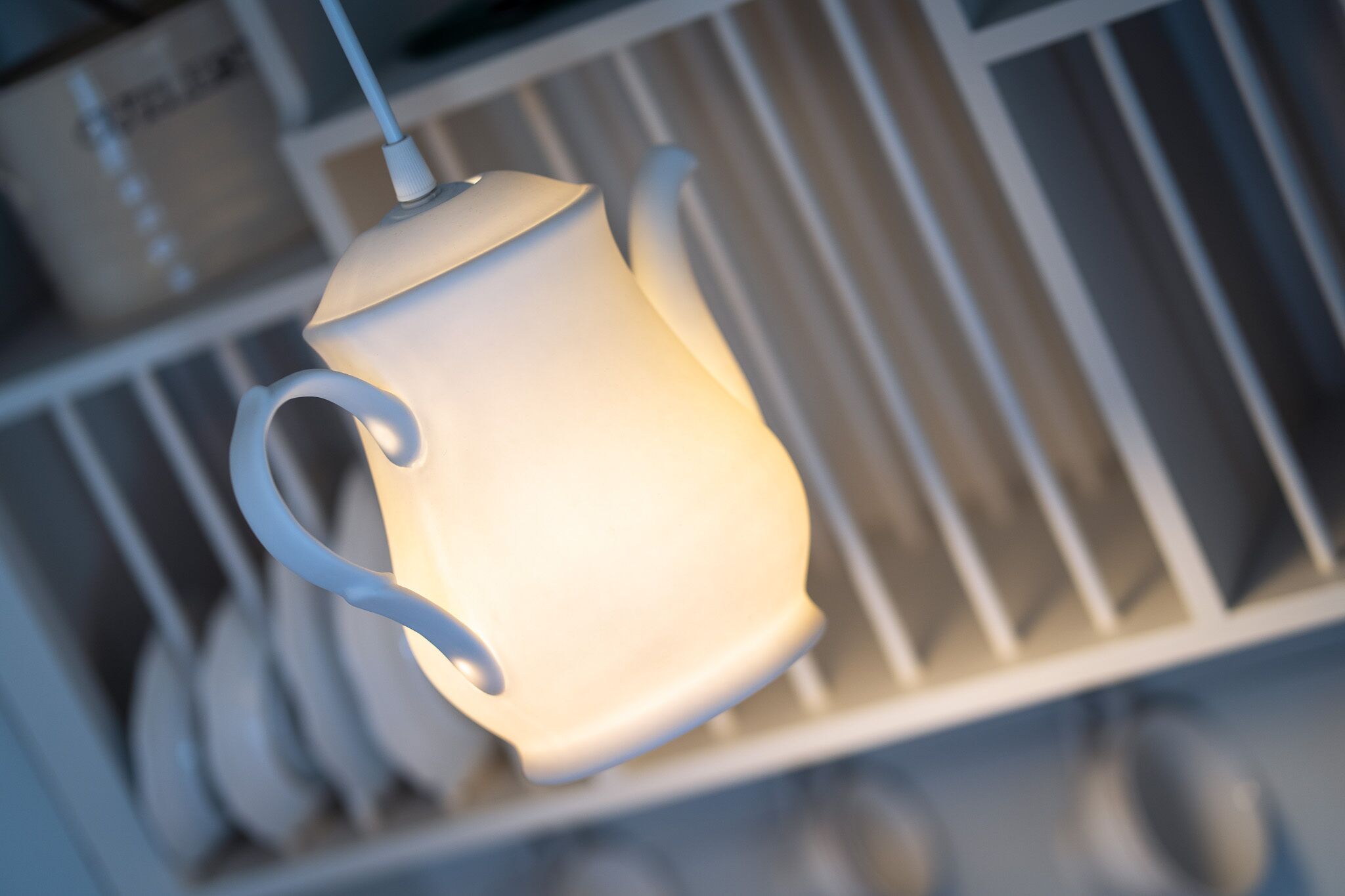 Eine weiße, teekannenartige Leuchte mit gebogenem Henkel, die von der Decke hängt.