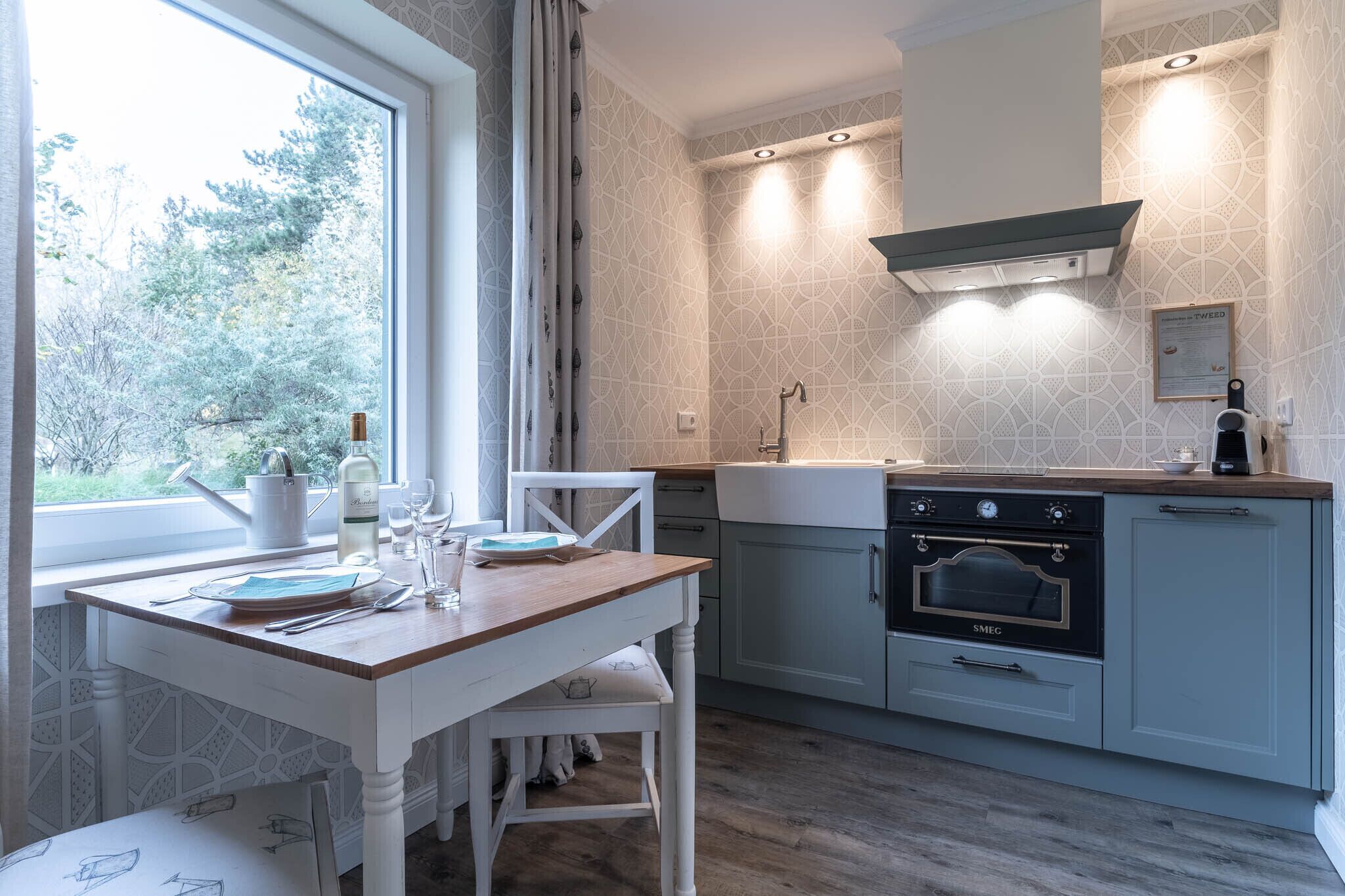 A modern kitchen with a wooden table, white chairs, a built-in oven, and a window overlooking greenery.