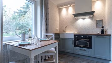 A modern kitchen with a wooden table, white chairs, a built-in oven, and a window overlooking greenery.