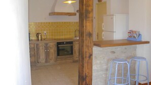 A kitchen with wooden cabinets, a stone countertop, a white refrigerator, and two blue bar stools. - Mas Las Vitalis (L'Isle-sur-la-Sorgue)