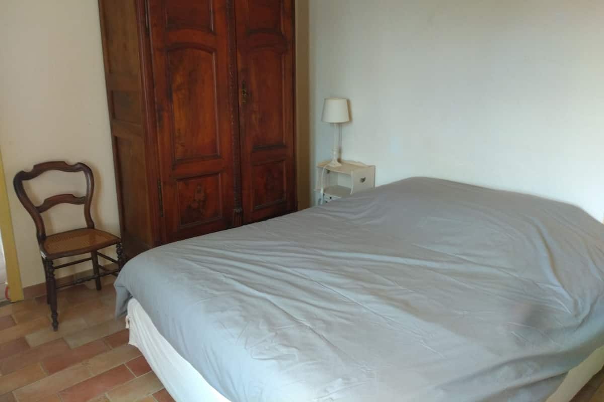 Un dormitorio con un armario de madera grande, una cama individual con colchón blanco, una mesita de noche con lámpara y una silla de madera.