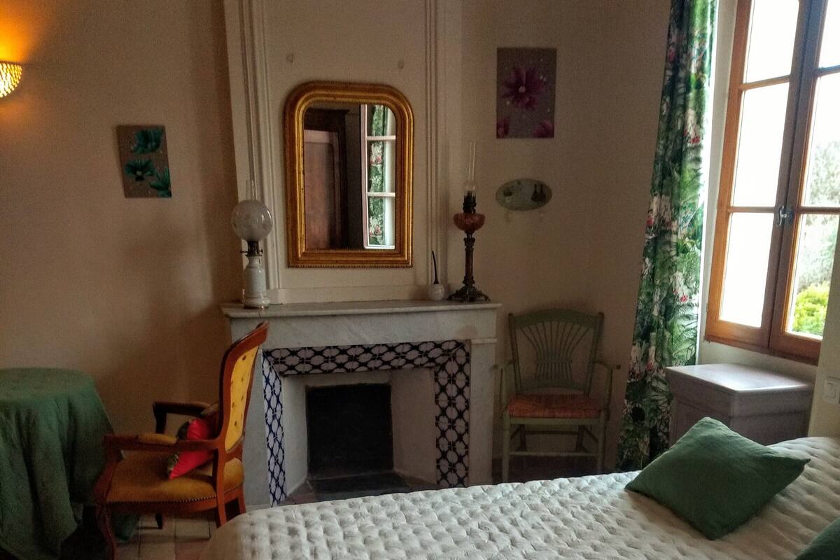 Un dormitorio con una cama grande, una silla, una mesa, una chimenea y una ventana con cortinas.