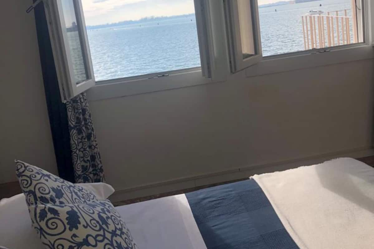 A hotel room with a bed, a window with a view of the water and a distant cityscape.