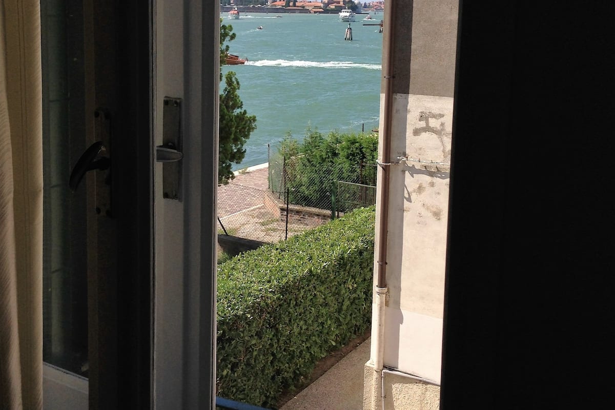 A view from a window overlooking a body of water with boats, a distant building, and a hedge.