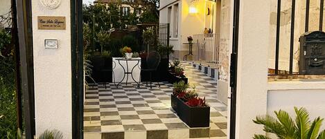 A checkered courtyard with a welcome mat, potted plants, and a table with chairs.