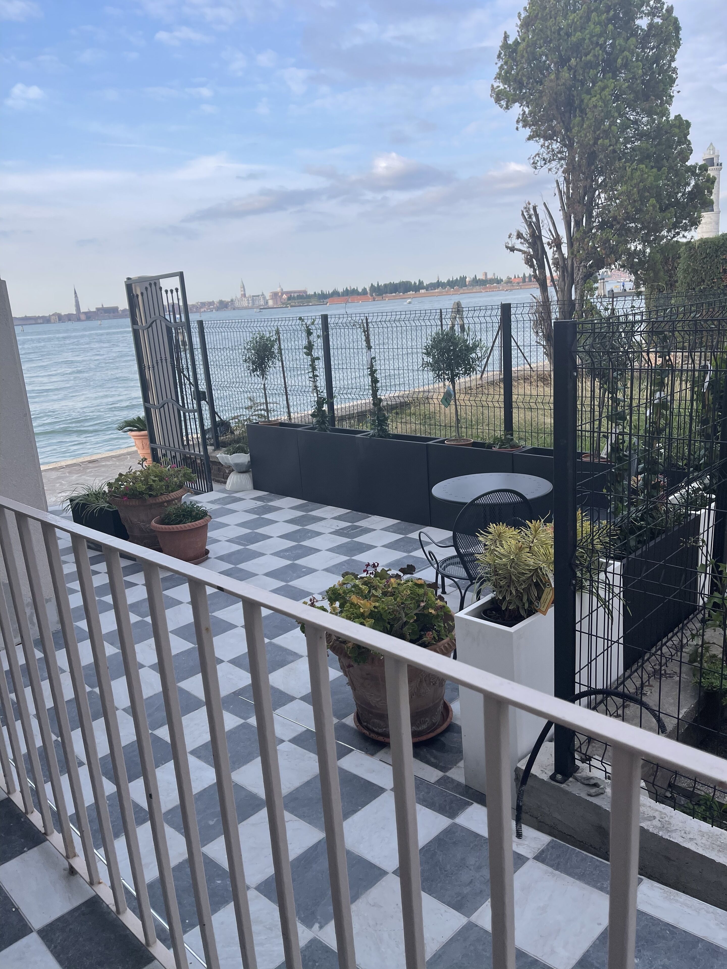 A balcony with a checkered floor, potted plants, and a view of the sea and distant buildings.