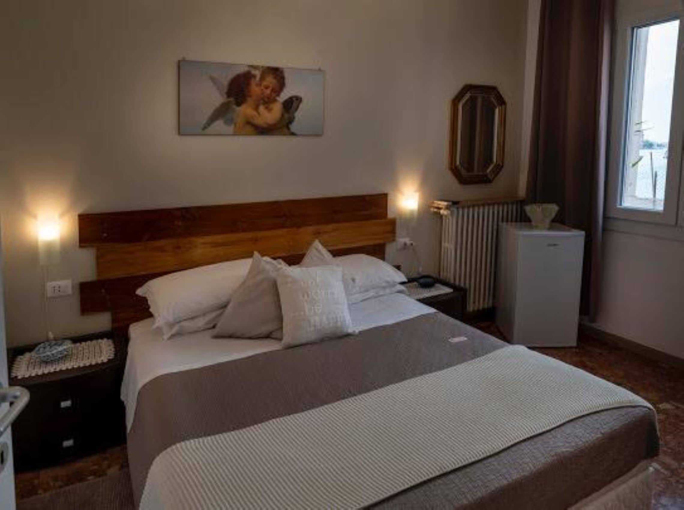 A hotel room with a wooden headboard, a bed with white linens, a nightstand with a lamp, a radiator, and a window with curtains.