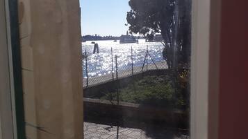 Blick durch ein Fenster auf eine pflasterbelagte Fläche mit einem Zaun, Bäumen und einem Gewässer.