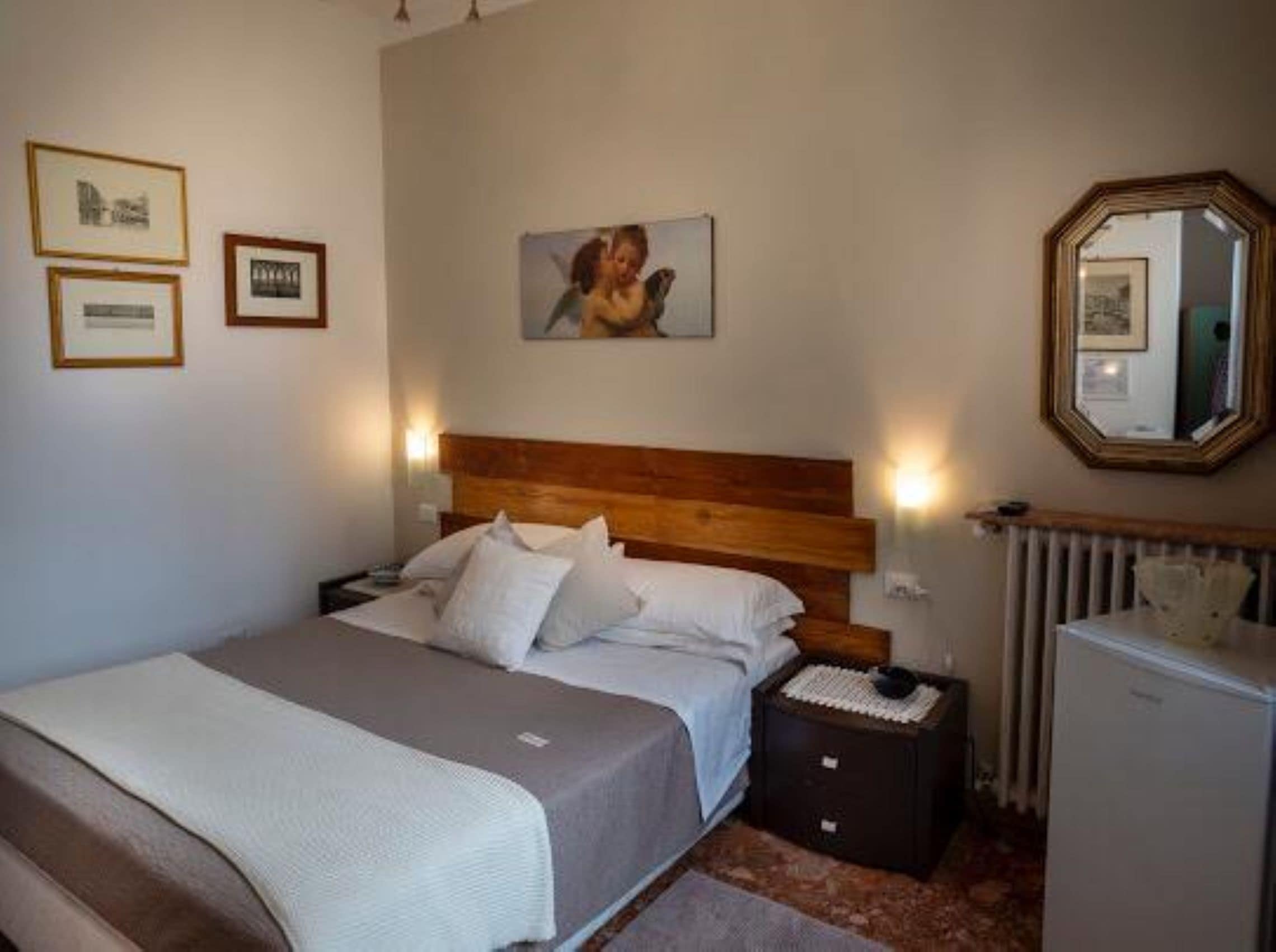 Una camera da letto con testiera in legno, un letto con lenzuola bianche, un comodino con una lampada, una cassettiera e uno specchio a parete.