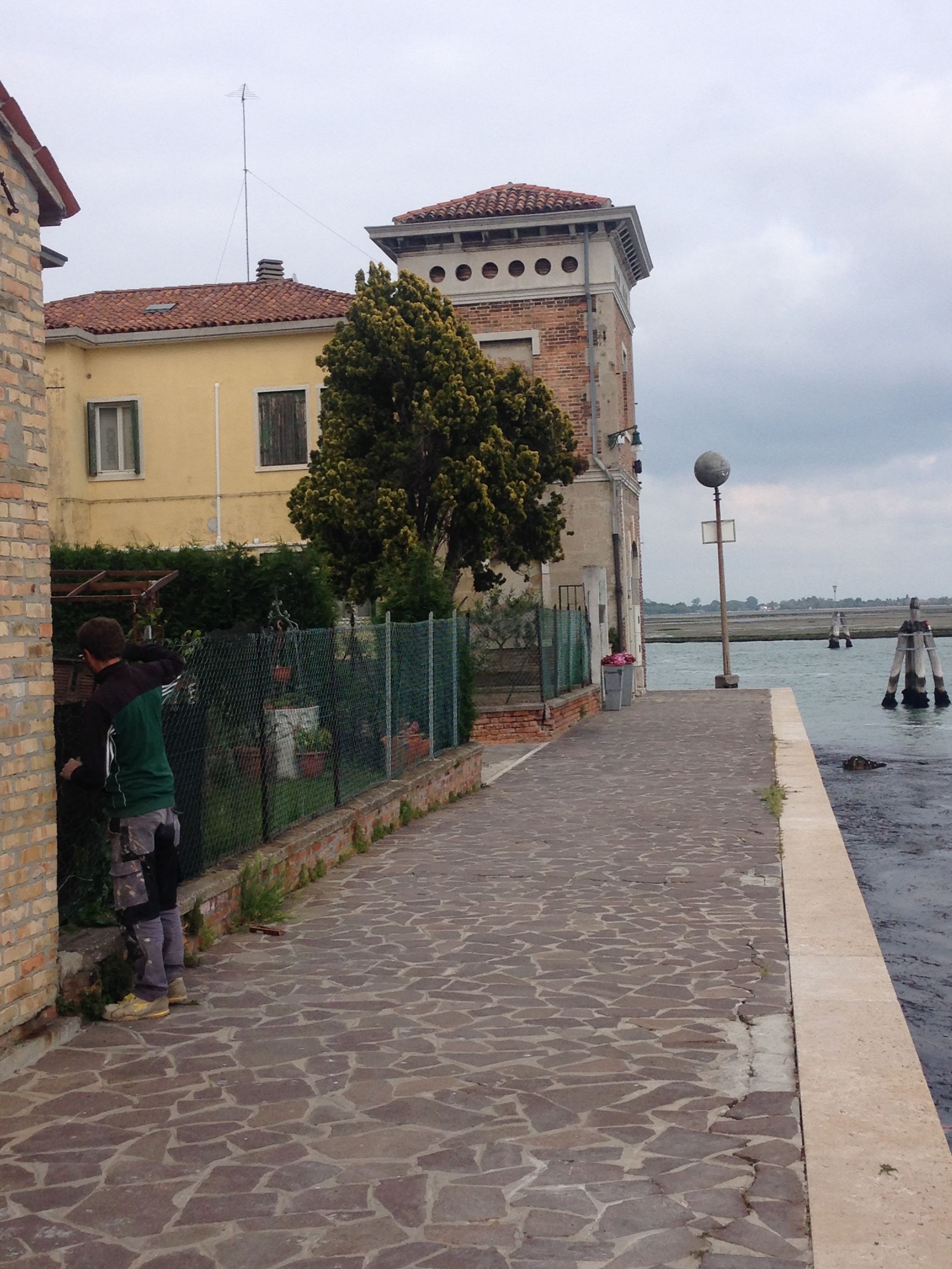Un chemin bordé de pierres, longeant un plan d’eau, avec une clôture et un bâtiment doté d’un toit en tuiles.