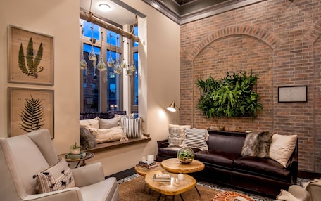 A cozy living room with a leather sofa, a white armchair, and a coffee table. There are framed botanical illustrations on the wall and a large window with hanging glass terrariums.