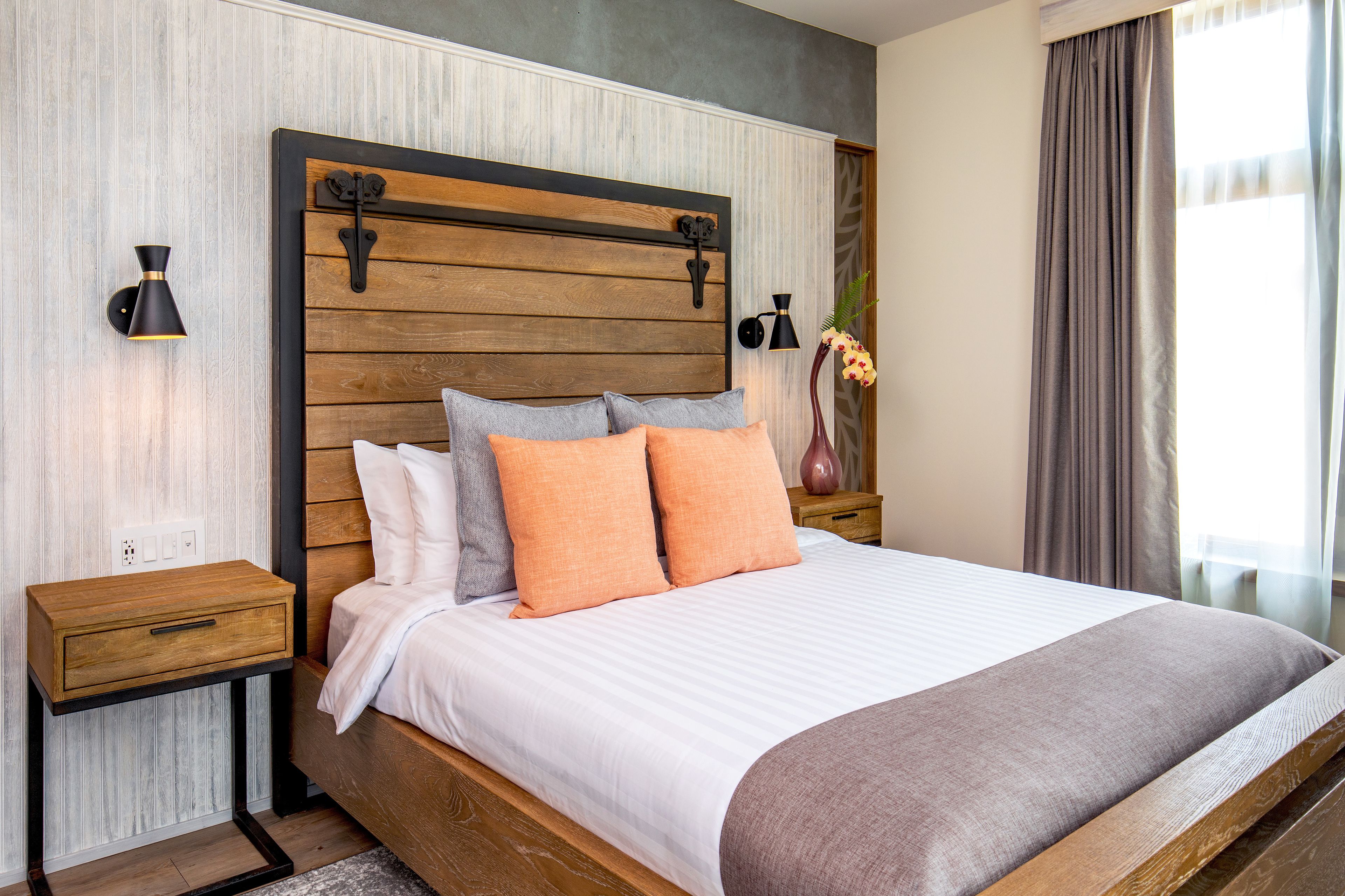 A modern bedroom with a wooden headboard, a large bed with white and orange bedding, two bedside tables with lamps, and a potted plant.