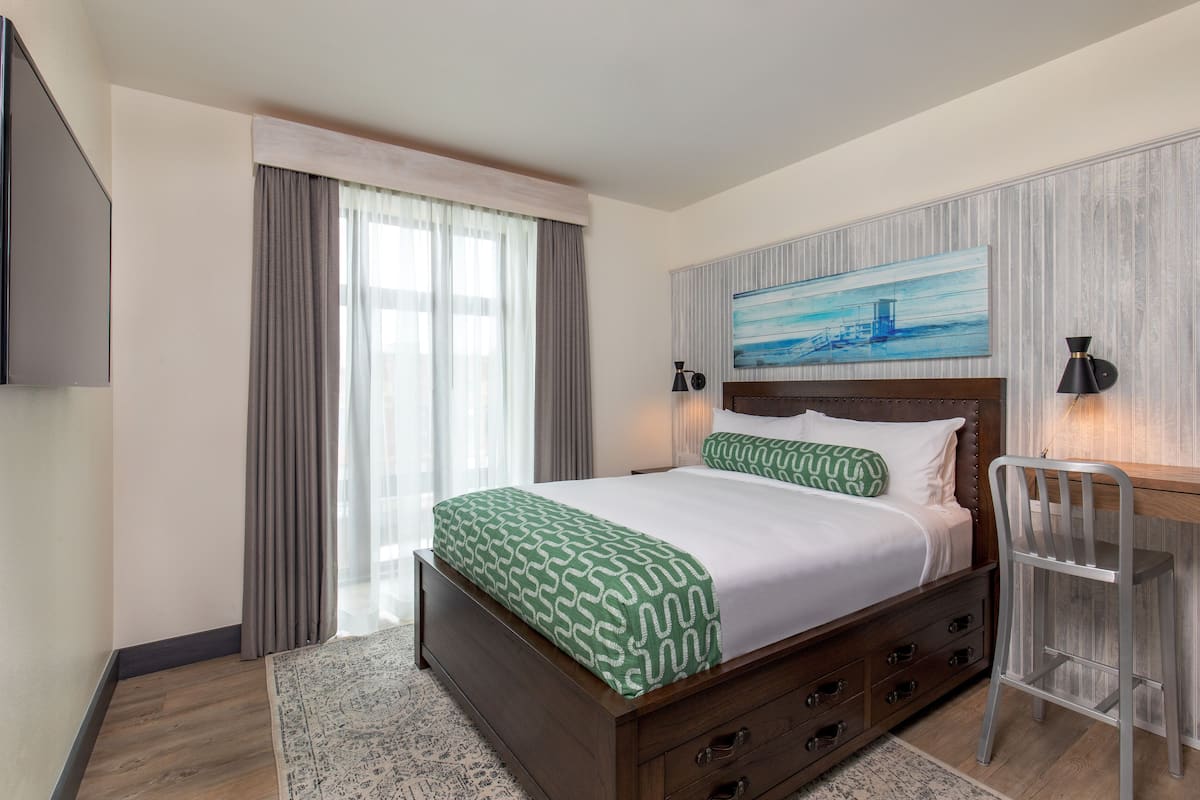 A hotel room with a bed, a desk, a chair, a TV, and a painting on the wall.