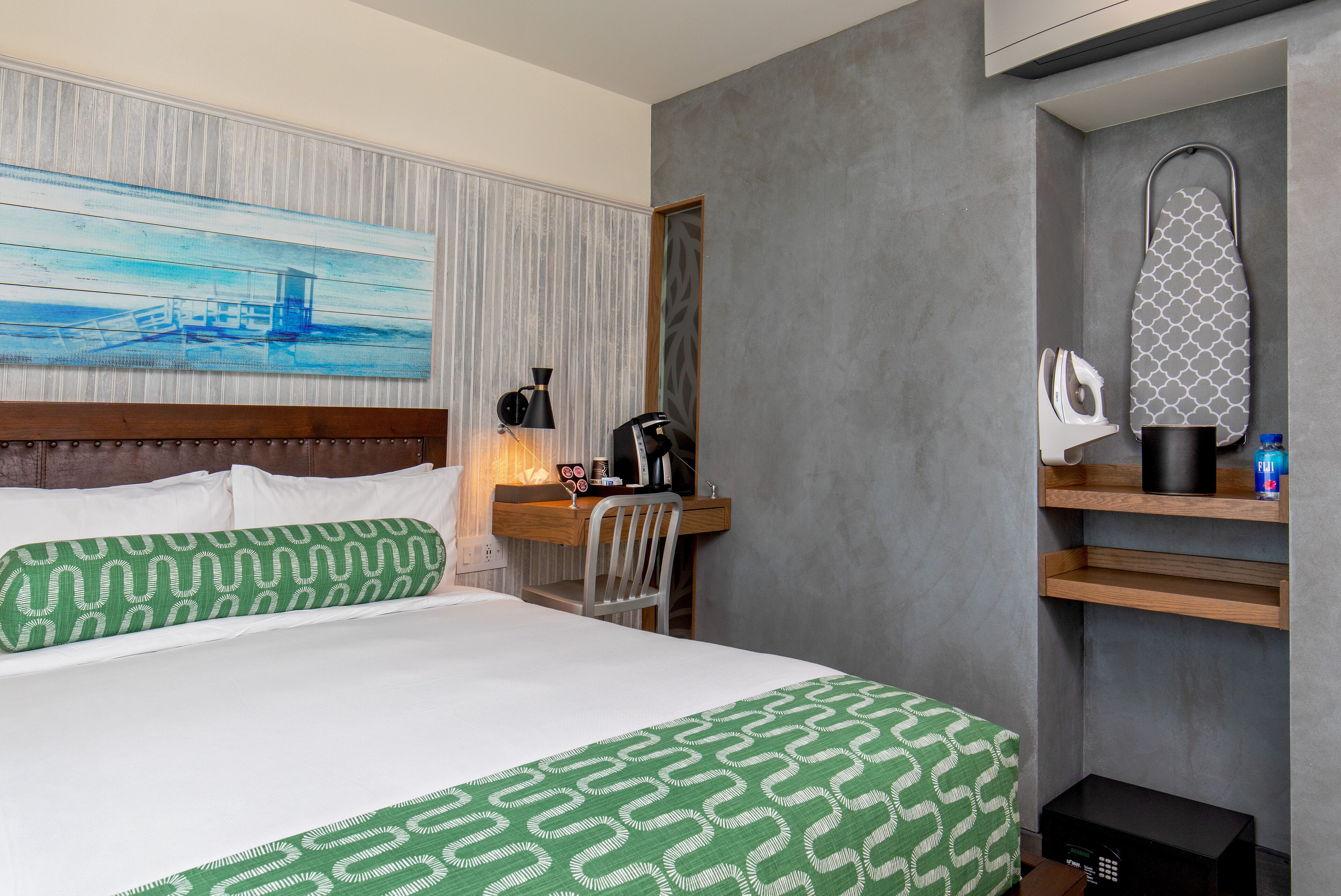 Ein Hotelzimmer mit einem Bett, einem Schreibtisch mit Stuhl, einer Kaffeemaschine, einem Bügelbrett und einem Bild an der Wand.