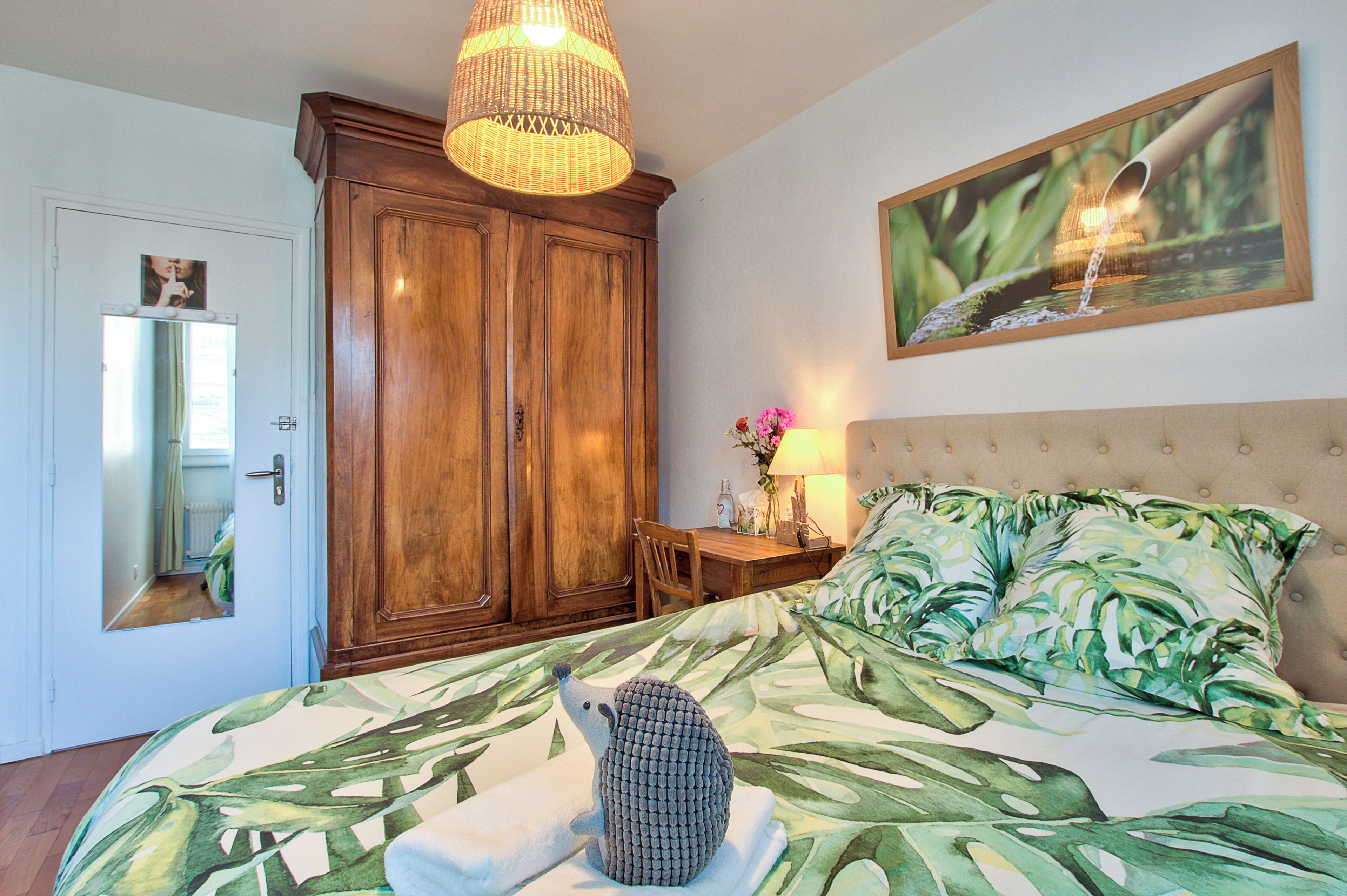 A bedroom with a bed, a wooden wardrobe, a mirror, a table with a lamp, and a painting on the wall.