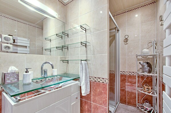 Un baño con lavamanos de vidrio, muebles blancos, un perchero y una zona de ducha con puerta de cristal.