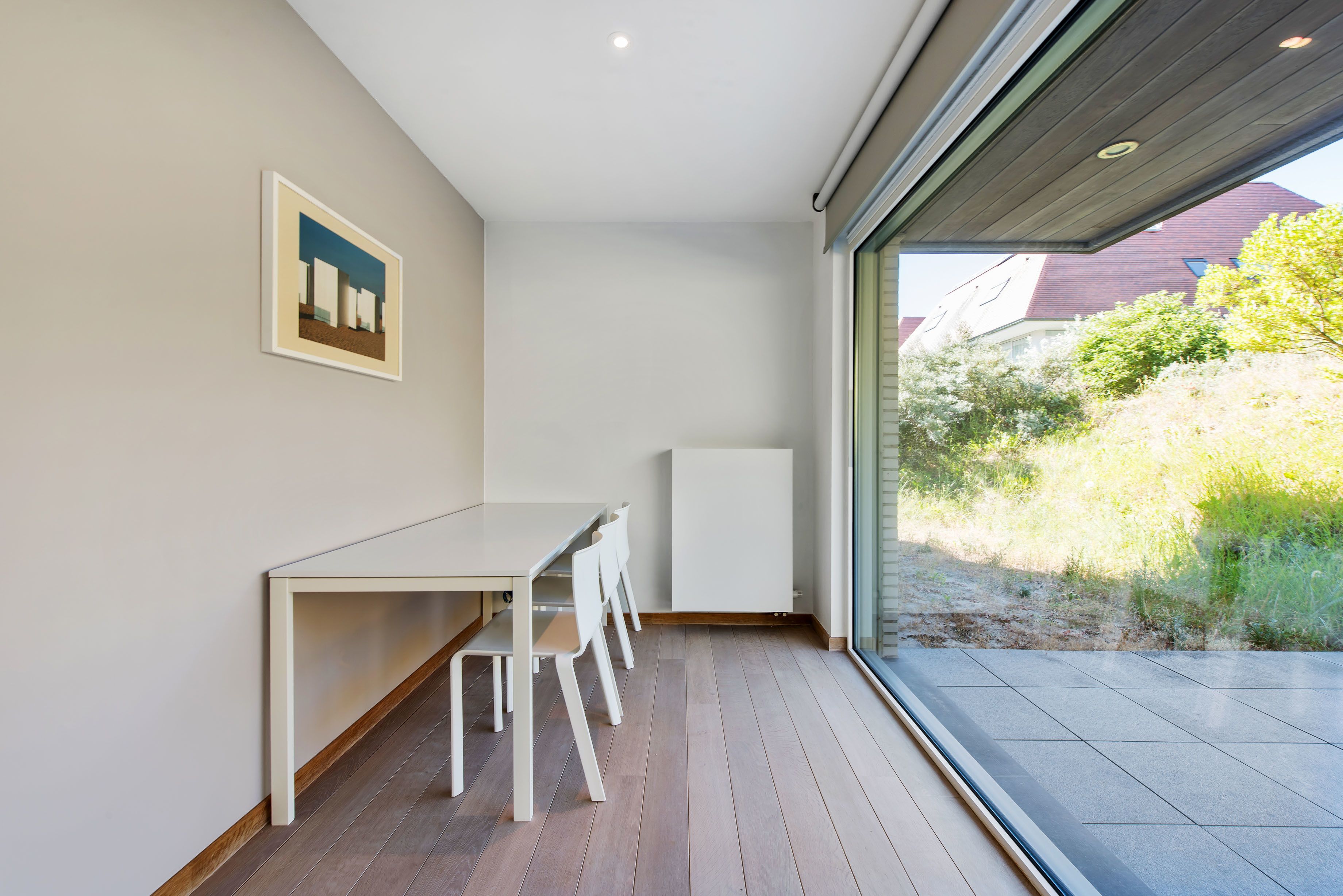 A modern interior with a white table and chairs, a framed picture on the wall, and a view of greenery outside.