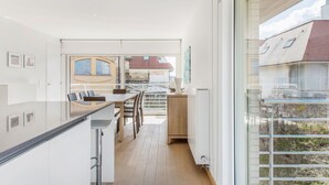 A modern kitchen with a central island, a dining area with chairs, and a view of the outside through large windows. - Zeegerucht Duplex (Koksijde)