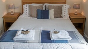 Cama bem arrumada com colcha listrada, dois travesseiros e duas toalhas dobradas no rodapé da cama. Há duas mesas de cabeceira com abajures de cada lado da cama.