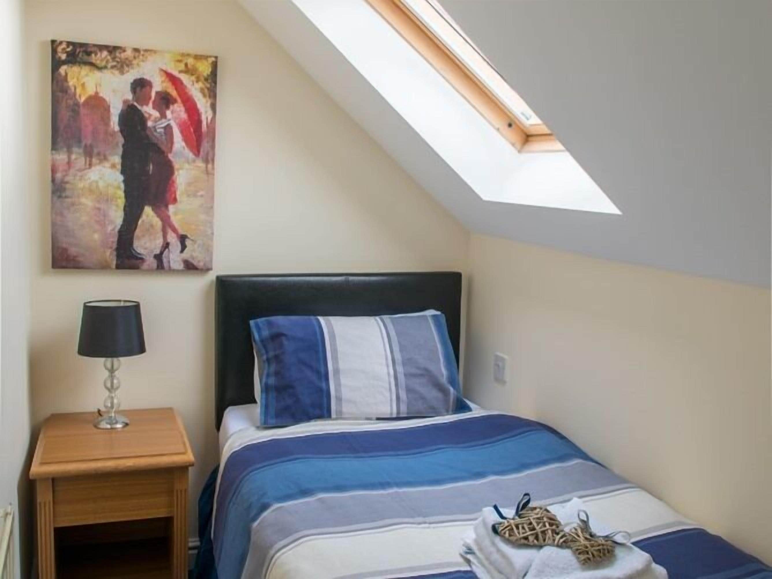 A bedroom with a sloped ceiling, a bed with a striped duvet, a nightstand with a lamp, and a painting on the wall.