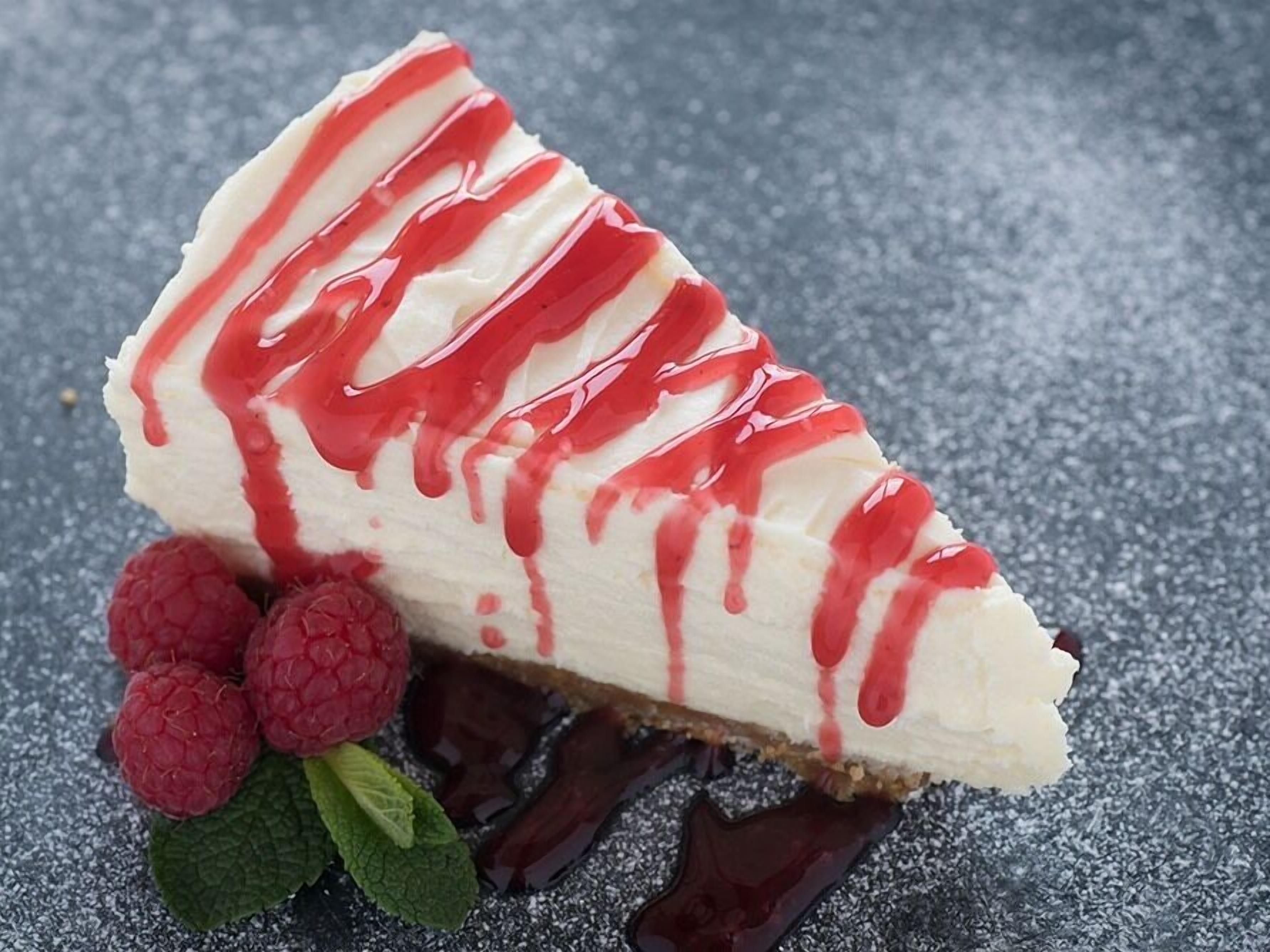 A slice of cheesecake with red syrup and raspberries on a textured surface.
