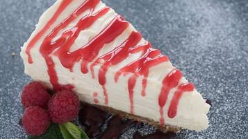A slice of cheesecake with red syrup and raspberries on a textured surface.