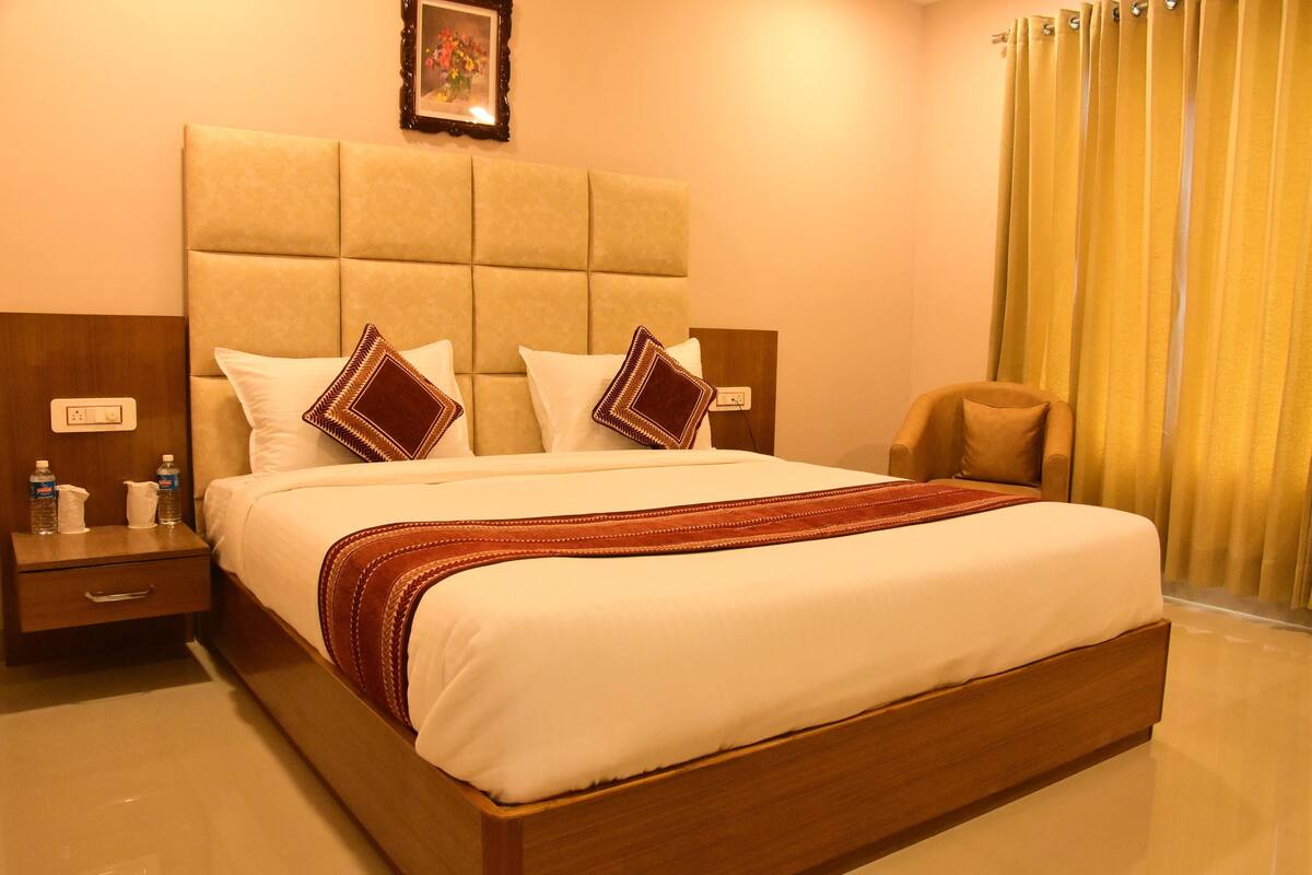 A hotel room with a large bed, a wooden nightstand, a chair, and a yellow curtain.