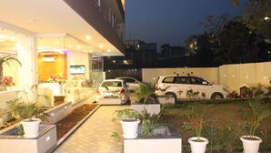 Entrada de hotel com balcão de recepção, plantas em vasos e carros estacionados.