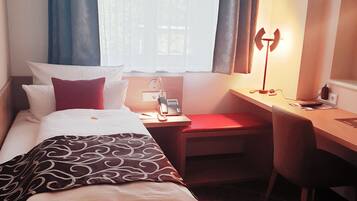 A hotel room with a bed, a desk, and a chair. There is a window with curtains, a lamp, and a phone on the desk.