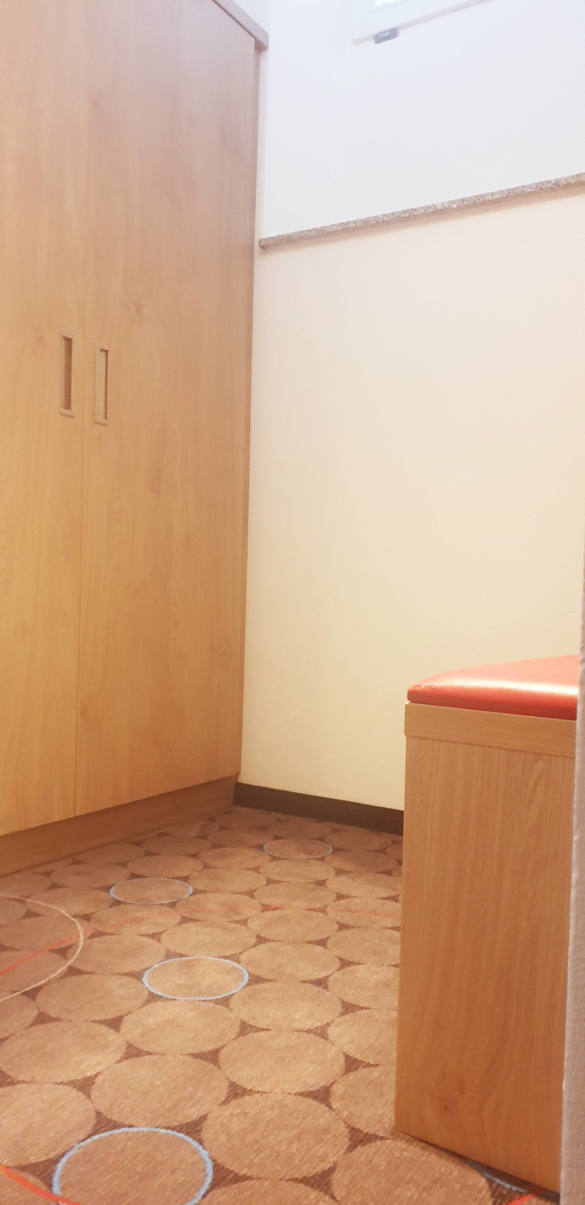 Een hoek van een kamer met een houten garderobe en een klein houten kastje met een rode bovenkant. De vloer heeft een vloertegel met een patroon.
