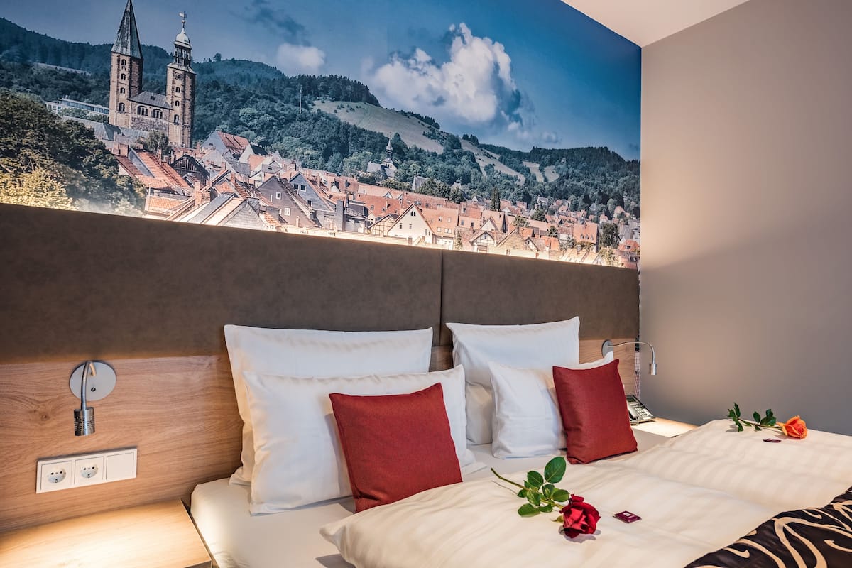 Ein Hotelzimmer mit einem großen Bett, einem Holzkopfteil und einem Wandbild einer Stadt mit einer Kirche und Hügeln.