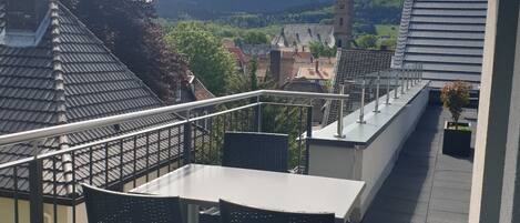A balcony with a table and chairs, overlooking a village with a church tower and hills.