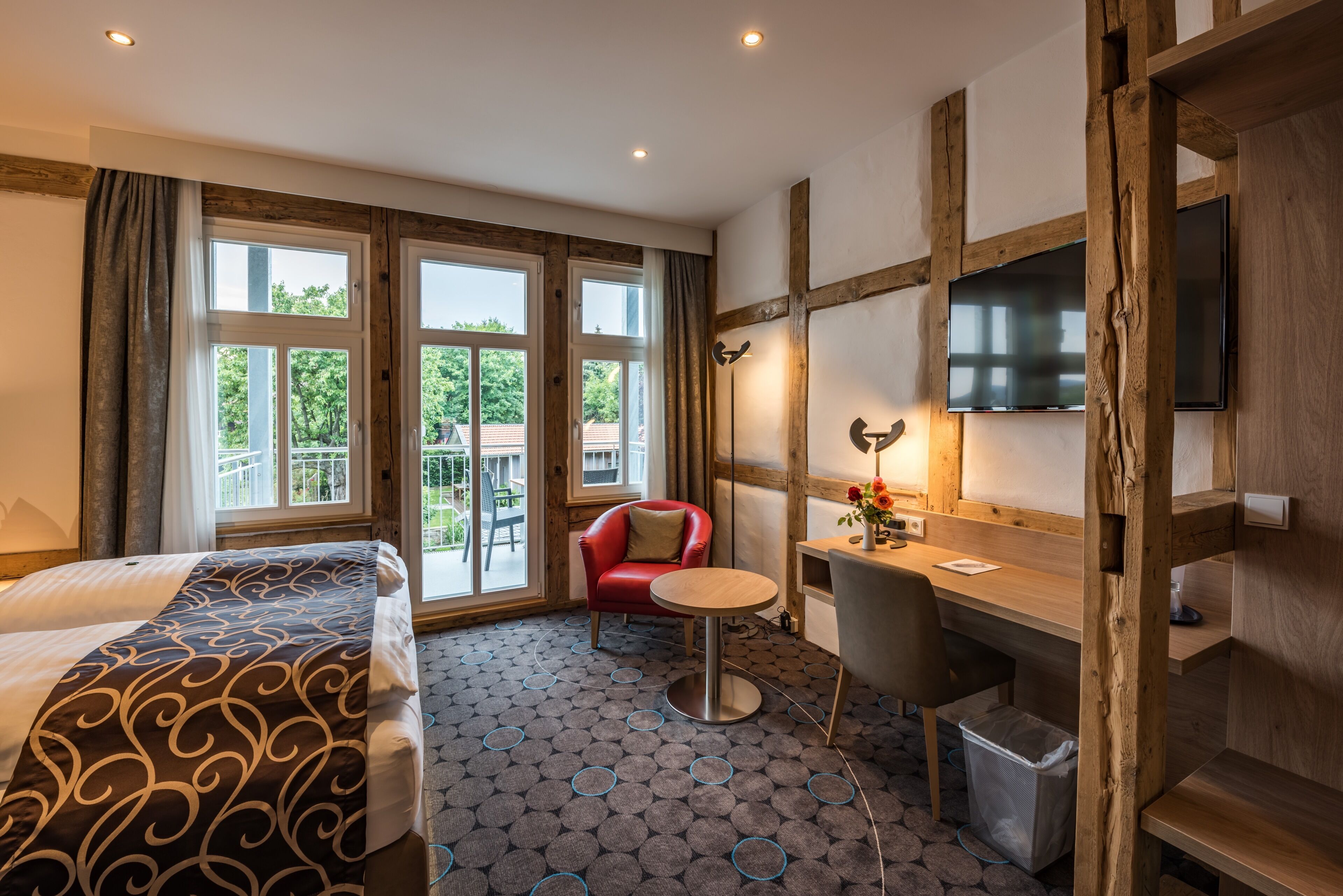 Una habitación de hotel con cama, escritorio, silla, televisor y ventanas amplias.