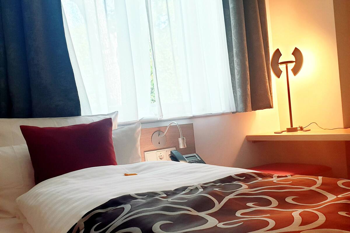 Ein Hotelzimmer mit Bett, Nachttisch mit Lampe, Fenster mit Vorhängen und einem an der Wand befestigten Telefon.