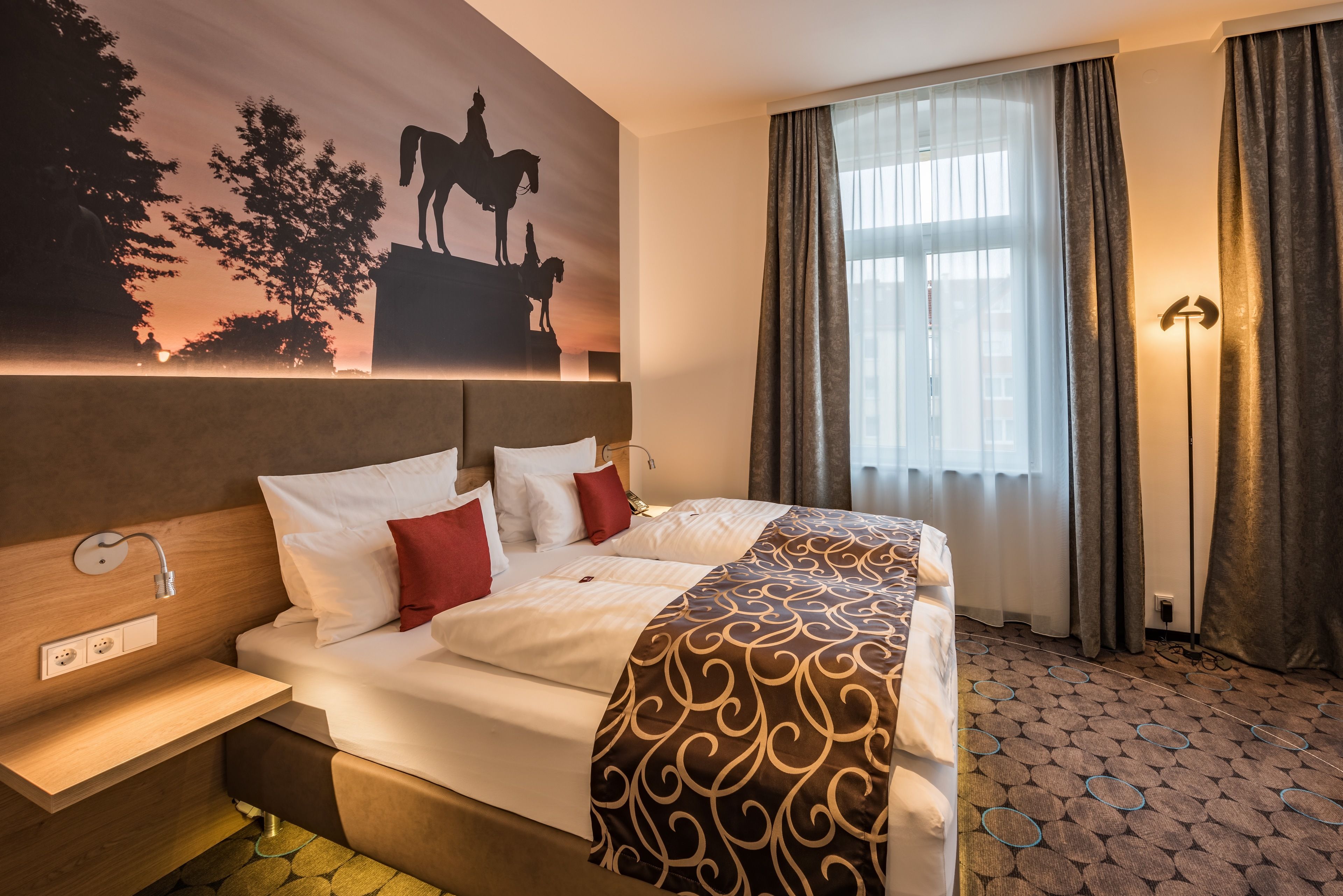 A hotel room with a large bed, a nightstand, a wall-mounted lamp, and a window with curtains.