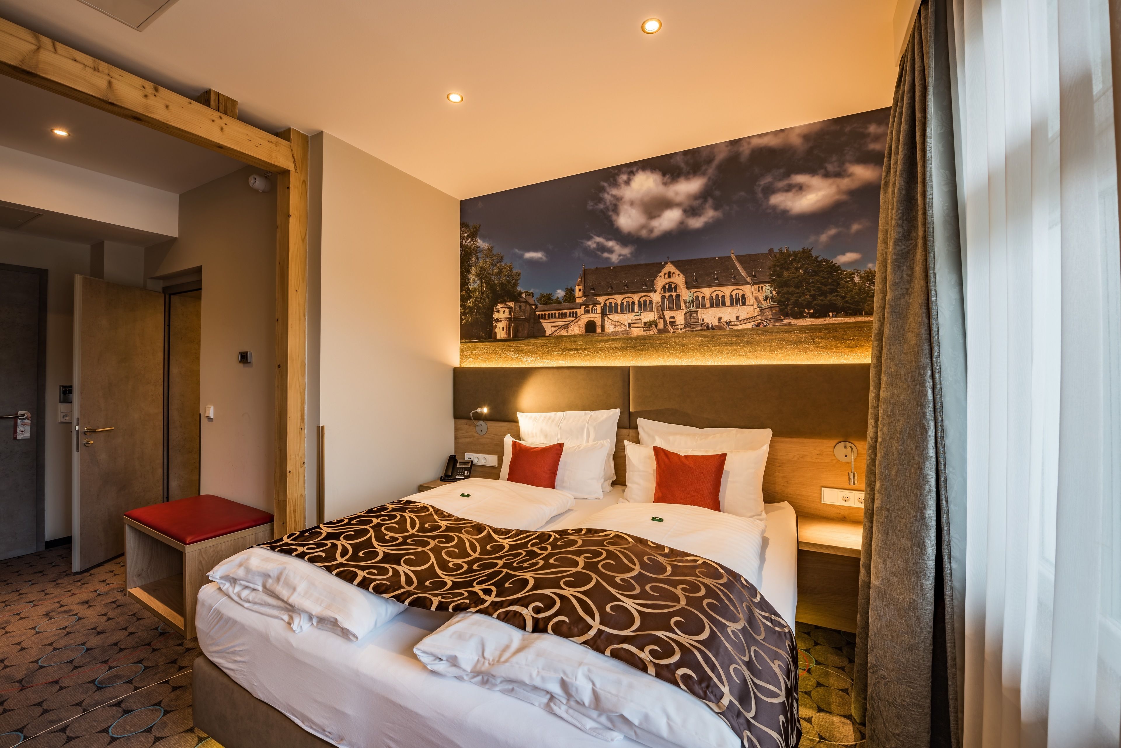Una habitación de hotel con una cama grande, un banco rojo, una mesita de noche y un televisor montado en la pared.