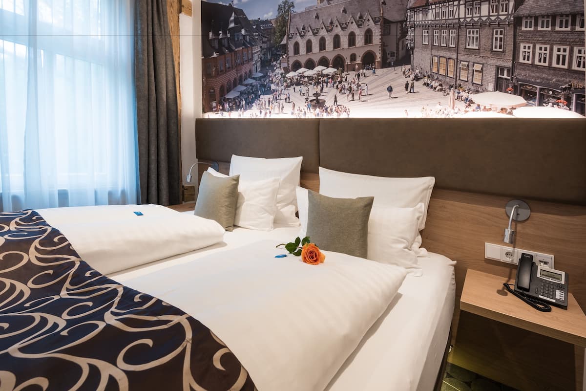 Ein Hotelzimmer mit zwei Betten, einem Holzkopfteil, einem Nachttisch mit Telefon und einem großen Fenster mit durchsichtigen Vorhängen.