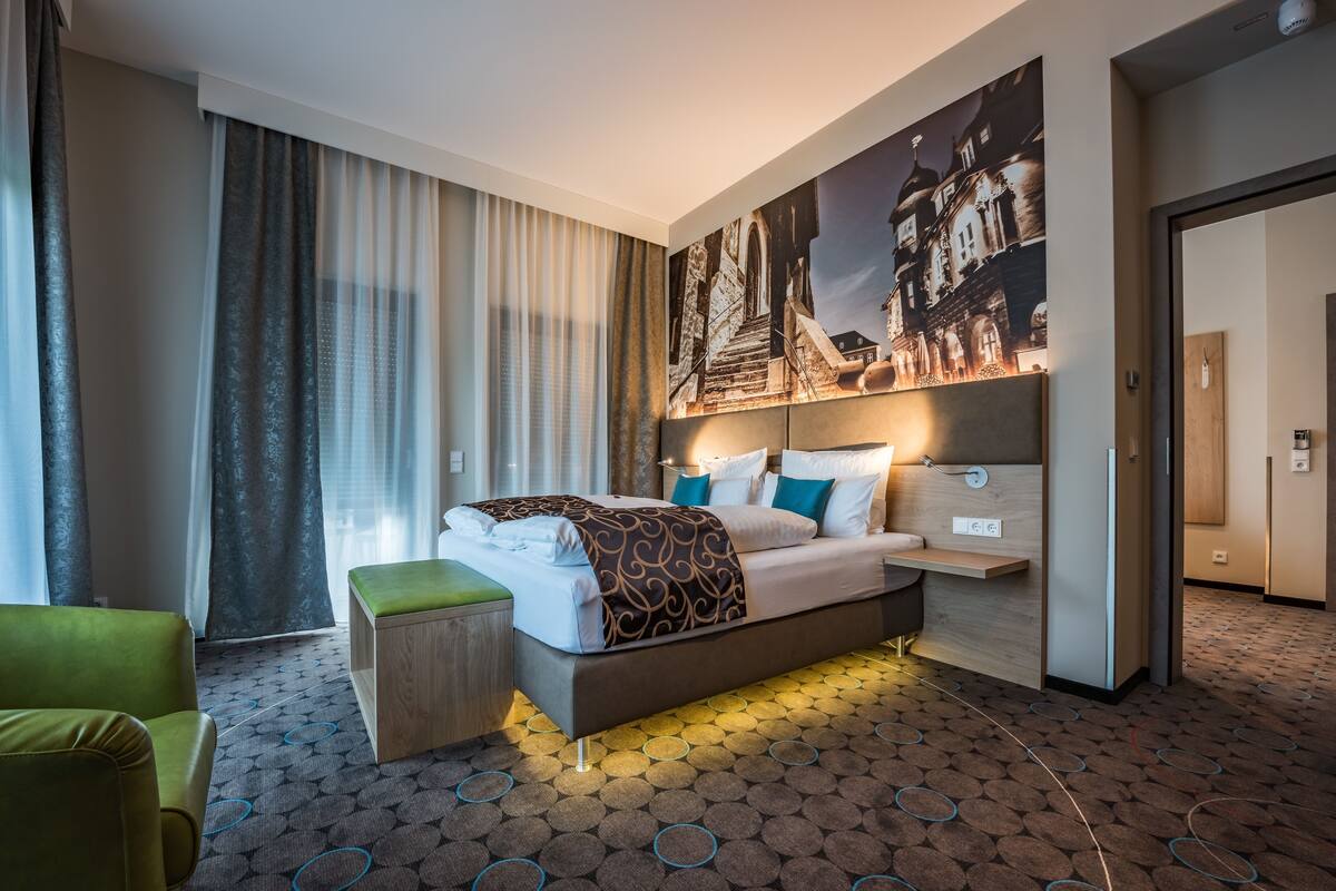 Ein modernes Hotelzimmer mit einem großen Bett, einem grünen Sessel, einem kleinen Holztisch und einem Stadtbild an der Wand.