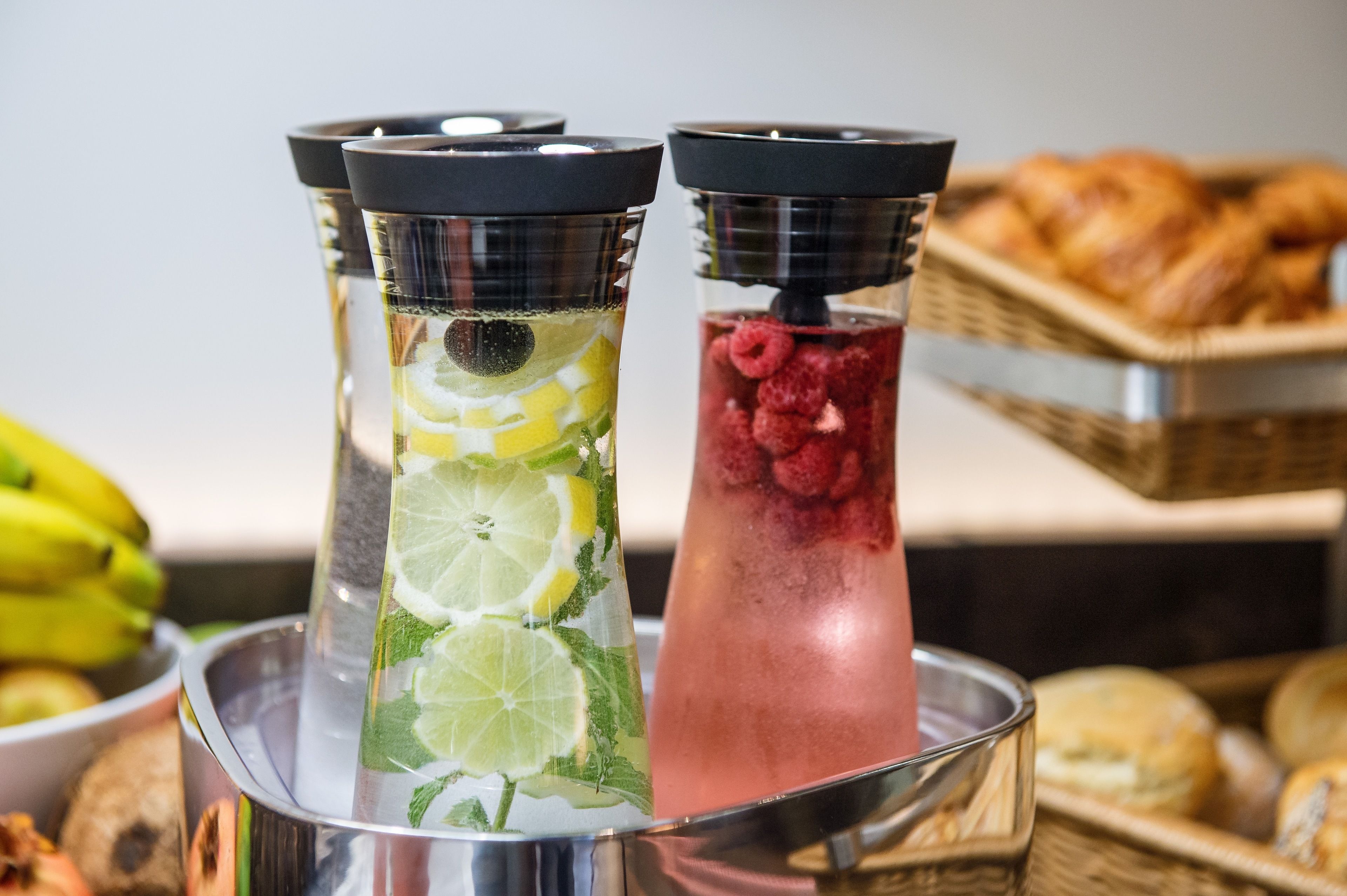 Two beverage dispensers with lime slices and mint leaves, and a basket of croissants in the background.