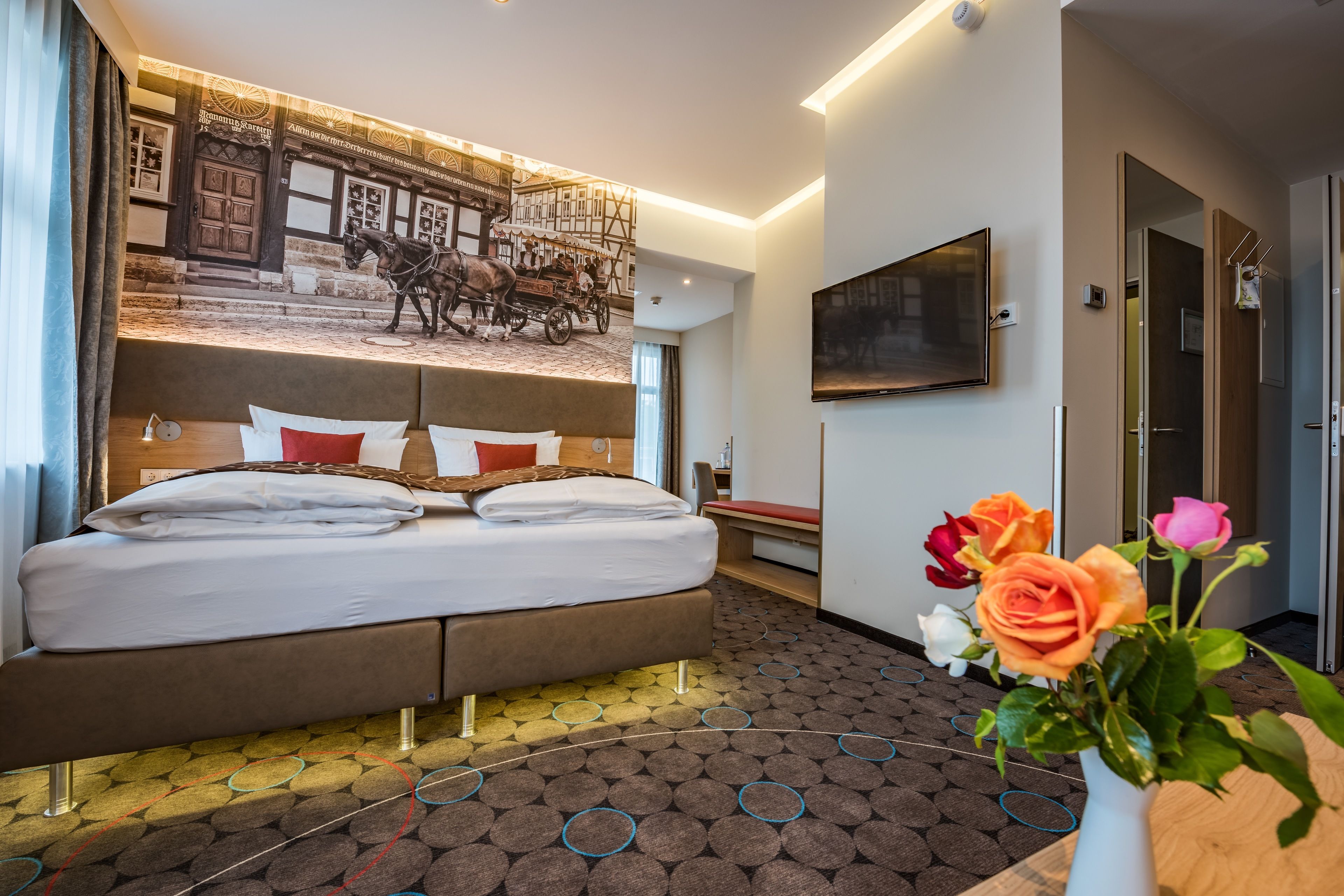 Quarto de hotel com uma cama grande, TV fixada na parede e um mural decorativo de uma carruagem puxada a cavalos.